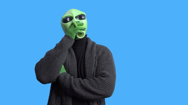 Professional actor in alien costume and cozy home outfit comes into the frame with funny mug and sits down to drink hot beverage against blue chromakey background...