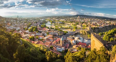 Aerial view from the Narikala fortress on the Tbilisi city, the capital of Georgia, full of small tiled roof houses, green trees, bridges over the Kura river in a sunny day.