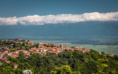 Fototapeta na wymiar Top view on a small city in Kakheti with many red-roof houses and green trees and the Alazani valley against foggy hills and clouds in a summer sunny day.