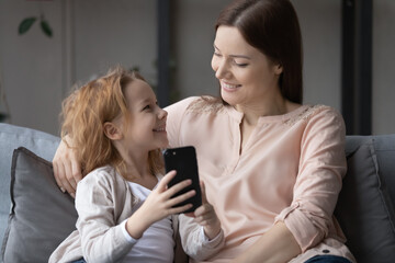 Fototapeta na wymiar Smiling cute little girl and young mother having fun with phone, sitting on cozy couch at home, happy family enjoying leisure time together, shopping online, watching video, browsing apps