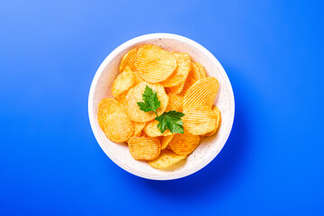 Fried corrugated golden potato chips with parsley leaf in wooden bowl on blue background, top view