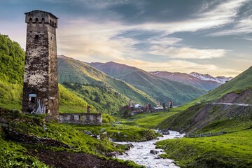 Obraz premium Gorgeous landscape in the Ushguli village, Georgia: medieval tower in the downhill on the bank of a rapid narrow river and green hills against pink sky in the background.