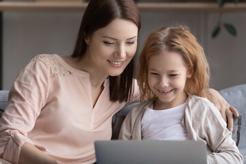 Close up smiling young woman with little girl using laptop at home, happy positive beautiful mother and adorable preschool daughter looking at computer screen, browsing apps, watching video