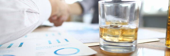 Close-up of glass with alcohol drink. Office workers making good deal. Partners celebrating signing important agreement and shake hands. Papers and documents on table. Business concept