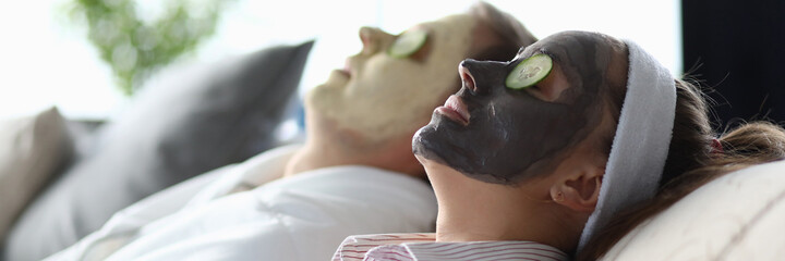 Naklejka premium Close-up of young woman face covered with clay moisturizing facial mask laying on couch with male friend. Beauty treatment for smooth and soft skin. Spa day and wellness concept
