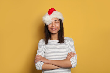 Happy black woman in Santa hat making a wish on yellow background