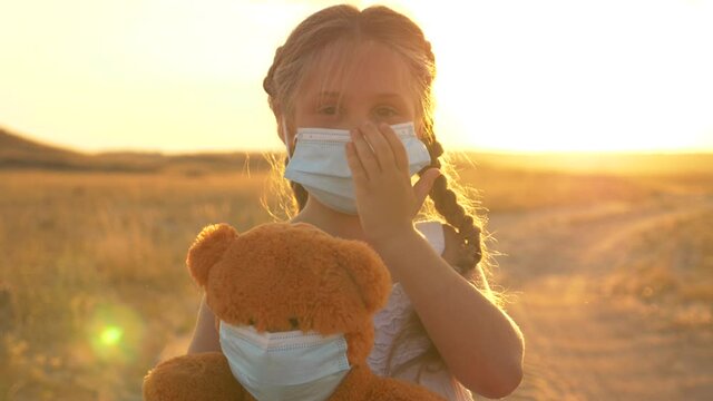 A child stands on the field with a soft toy in his hands. A child in a medical mask isolated during the quarantine of the virus. A kid plays doctor with a Teddy bear. The concept of health care.