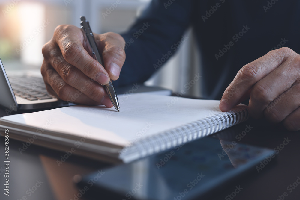 Poster close up of man hand writing on paper notebook with a pen while working on laptop computer