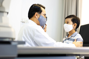Fototapeta premium An Asian male doctor uses An ophthalmoscope to examine the boy's eyes. The ophthalmologist examines the child's vision. They wear a protective face mask.
