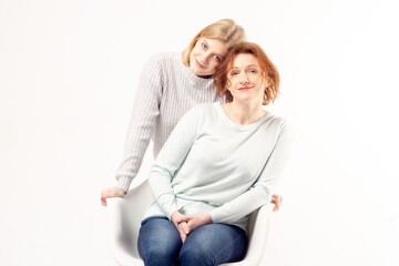 Young teenage blonde girl hugging her smiling ginger hair senior mother sitting on the chair