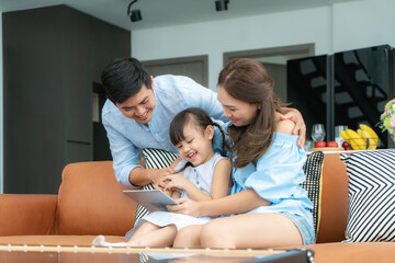 Happy Asian family with father, mother and daughter sitting on sofa and using digital tablet looking at screen together in living room at home..