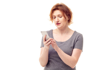 Red haired adult mature woman in gray dress looking on mobile phone in her hands with pensive expression