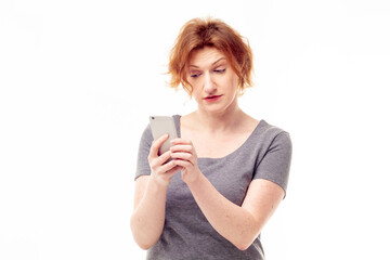 Red haired adult mature woman in gray dress looking on mobile phone in her hands with pensive expression