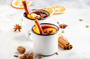 Mulled wine in white metal mugs with cinnamon, spices and orange on gray background, traditional...