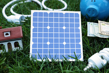 Composition with solar panel, house model and lightbulb on green grass