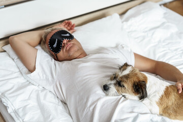 Mann über 50 mit einer Augenmaske und ein kleiner Terrier Hund schlafen auf einem Bett....