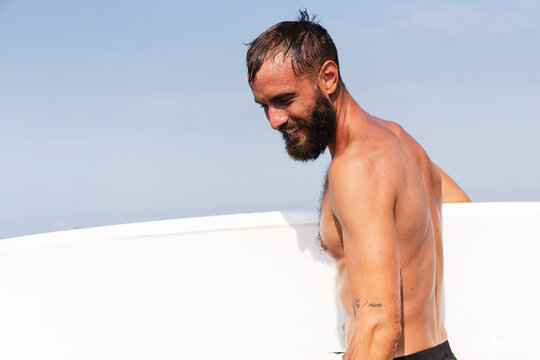 Young smiling surfer holding his surfboard on the beach - Handsome hipster man waiting waves for surfing - Sporty bearded man training with surfboard to sea - Lifestyle and freedom concept - Copyspace