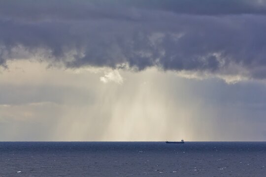 A cargo ship floating afar in the Baltic sea against cloudy sky and a sunbeam. © Алексей Мараховец