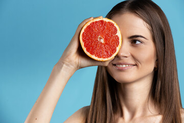 Pretty caucasian woman holding juicy half of grapefruit against her eyes, making gorgeous face. Blue isolated background