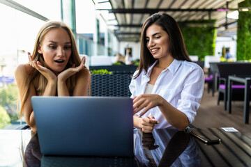 Two young women, bloggers, wearing in shirts are sitting in cafe at table and using laptop, working, blogging, online shopping.