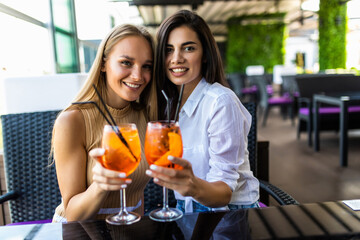 Young women drinking aperol spritz, clink glasses with friends, looking at camera in modern cafe