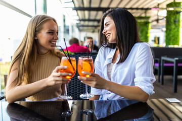 Blissful pretty women sitting near to big window with glasses of icy cocktails and laughing.