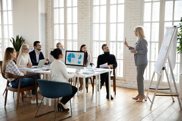 Obraz premium Confident female speaker or coach stand make whiteboard presentation at meeting with diverse employees. Successful woman trainer talk present business project on flip chart for workers in office.