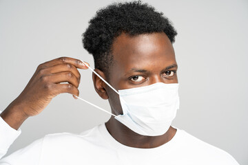 Studio portrait of African American young man  wearing face medical mask, looking at camera, isolated on grey background. Protection against flu, virus, Covid-19, coronavirus pandemic. New normal.