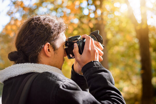 Young professional male photographer takes pictures in the autumn park. Golden trees and bright sun on the background.