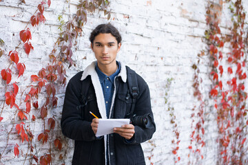 Portrait of handsome beginner photographer standing near white wall, holding copybook and pencil in...
