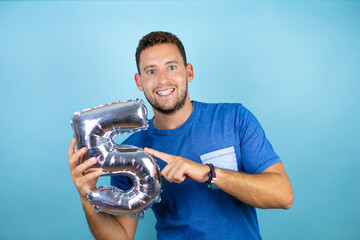Fototapeta premium Young handsome man wearing blue casual t-shirt over isolated blue background holding a number five balloon. Celebration concept