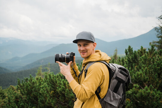 Young male traveler photographer. Attractive man photographer in hiking wear stands in the mountains holding a camera in his hands, smiles and looks at the camera against the backdrop of a mountains
