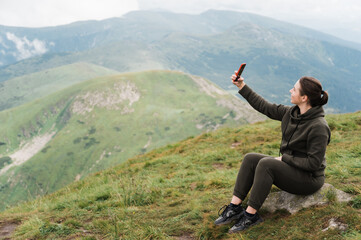 Caucasian happy young woman climbing the top of the mountain and taking a selfie on the phone while sitting on the stone against the background of an amazing mountain landscape