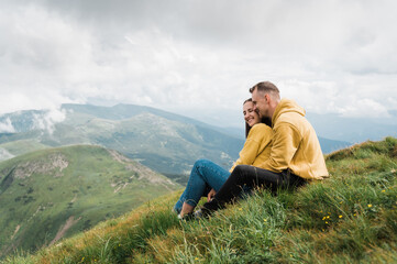 Fototapeta na wymiar Happy couple in love hugs and rests while sitting on the top of the mountain after a hard climb watching a beautiful mountain landscape