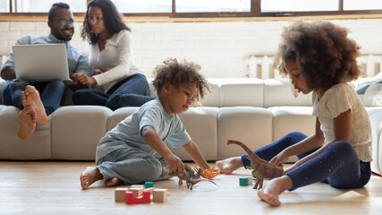 Cheerful adorable little mixed race children siblings playing toys on warm wooden floor, while young couple parents relaxing on comfortable sofa, web surfing on computer, happy family weekend.