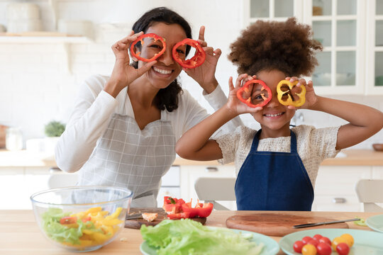 Happy african american family in aprons making funny grimaces with pepper, looking at camera. Portrait of joyful mixed race mother having fun with adorable pretty little kid daughter in modern kitchen - Powered by Adobe