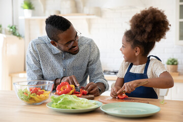 Joyful african american man in glasses having fun with adorable biracial kid daughter, preparing healthy food together in modern kitchen, happy mixed race family enjoying chopping vegetables for salad