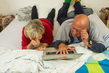 elderly husband and wife on the bed in their bedroom talking and laughing and having fun watching the laptop computer passing joyful moments