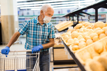 older european man wearing mask and gloves with covid protection chooses buns and bread in supermarket bakery