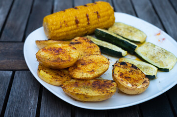 vegetarian barbecue plate with zuchini, corn on the cob and potato halves with spices on a wooden table