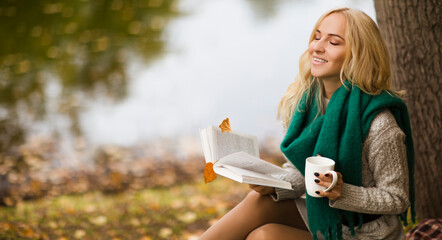 Beautiful young blonde woman sitting on a fallen autumn leaves in a park, reading a book and drinks tea
