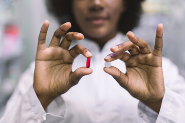 Young smiling African woman doctor or clinical pharmacist holding white white tablet pill and red...