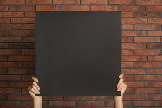 Woman holding black blank poster near red brick wall, closeup. Mockup for design