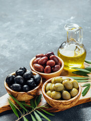 Set of green, black and red or pink olives and olive oil on gray background. Different types of olives in olive wooden bowls and olive oil over wooden cutting board. Copy space. Vertical