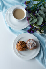On a white-blue background, a bouquet with flowers, a cup of coffee with milk and a plate with cupcakes.