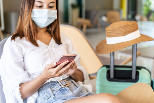 Young Woman Wearing Medical Masks With Baggage Using Smartphone While Waiting To Check In The Hotel, New Normal Travel Lifestyle Concept