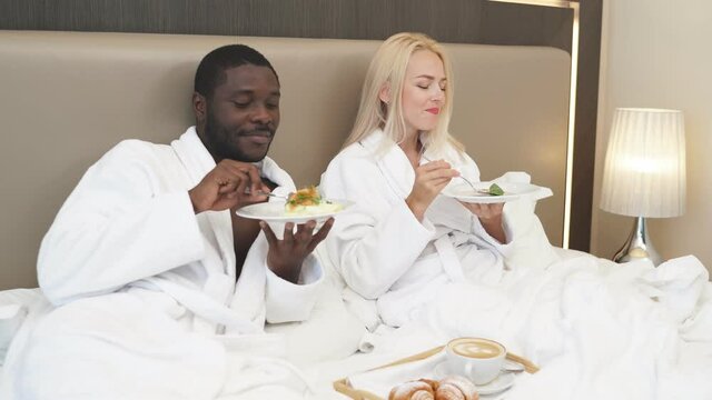 Adorable multiethnic couple have breakfast in hotel, they have tasty meal together, enjoy, relax, happy weekends. on bed.