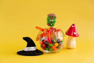 Lollipop in the form of a green monster in a round jar with multi-colored sweets on a yellow background.