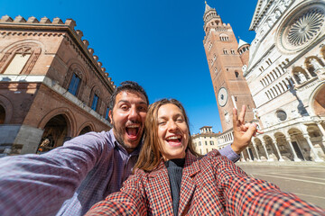 happy tourist couple visiting the city of Cremona taking selfie in the middle of main square of Cremona with Cathedral, baptistery and Torrazzo bell tower . 