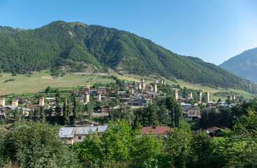 Fototapeta na wymiar Panoramic view of Mestia, a highland townlet in Georgia in the Caucasus Mountains. Houses with stone defensive towers on the foreground and surrounding mountains with green forest on the background.